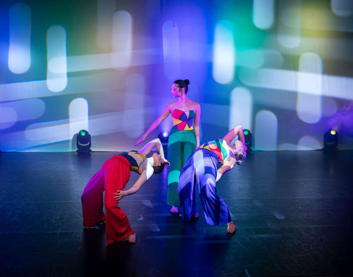 Three dancers on a dark stage under blue lighting; two are bending backwards and one is standing straight with one arm extended
